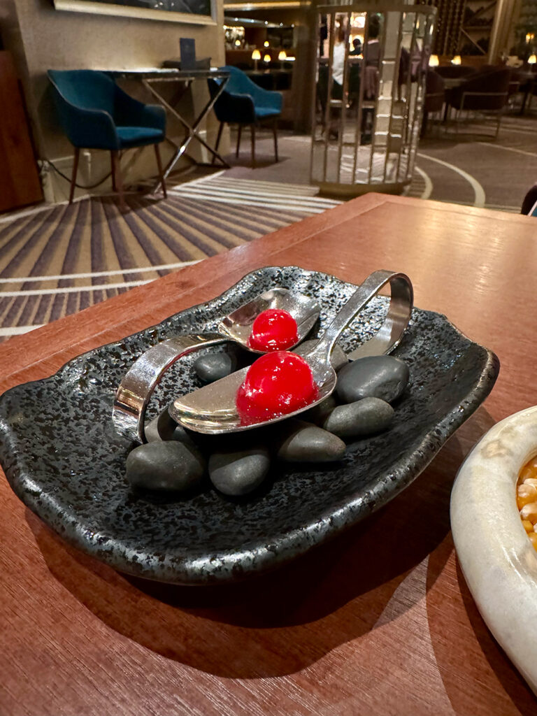 two spoons sit on a black slate, with red spheres sitting in the spoons.