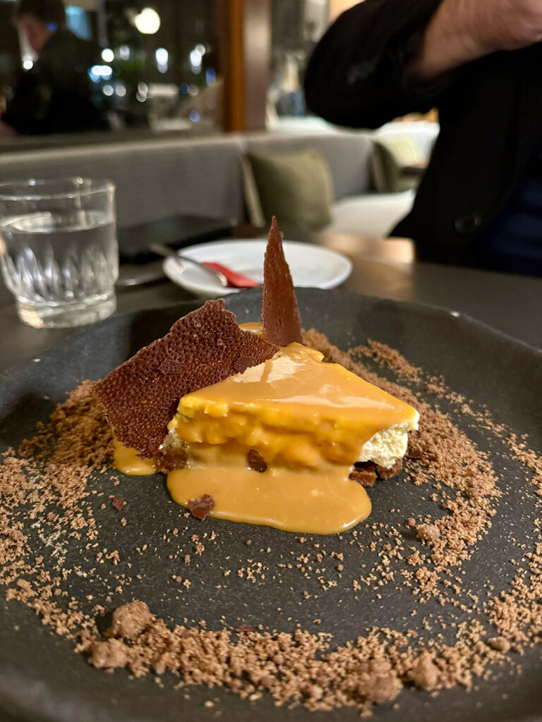 A slice of cheescake covered in salted caramel sauce sits on a black plate surrounded by crumbled biscuits. 