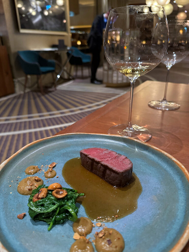 a slice of eye fillet sits on a blue plate with wilted spinach, hazlenuts and leek puree accompanying. 