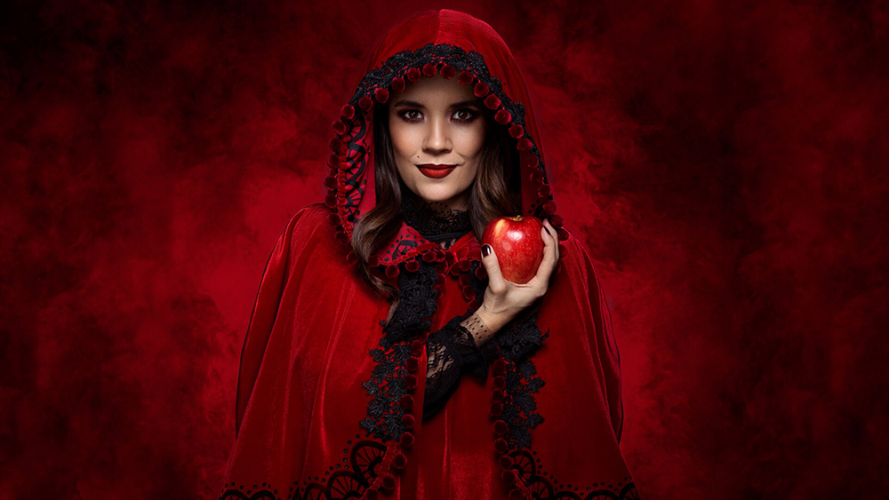 A brunette in a blood red cape holds out an apple.