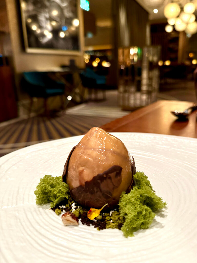 A dessert that looks like a giant hazelnut sitting on a forrest floor sits on a white plate.