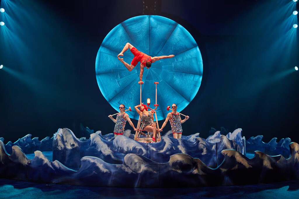 Circus performers in bathing suits perform in front of a seaside theatre set. 