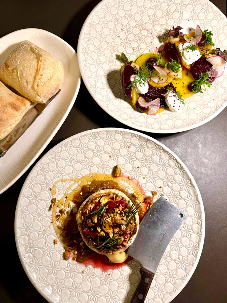 Top down photo of woodfired camembert topped with pomegranate and a plate of assorted colourful beets. A plate of bread is off to the side. 