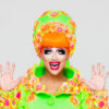 Bianca Del Rio smiles open mouthed, with both her hands up. She is wearing neon green and has a neon orange wig.