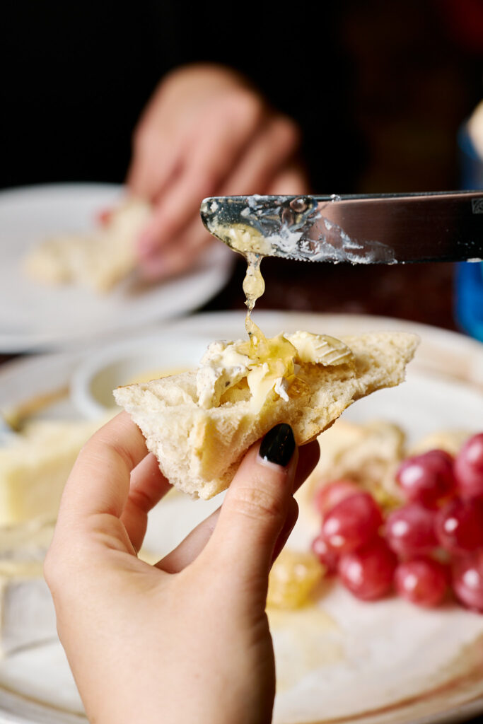 A hand holds a wedge of baguette with truffled brie on top, while a knife drizzles honey over it. 