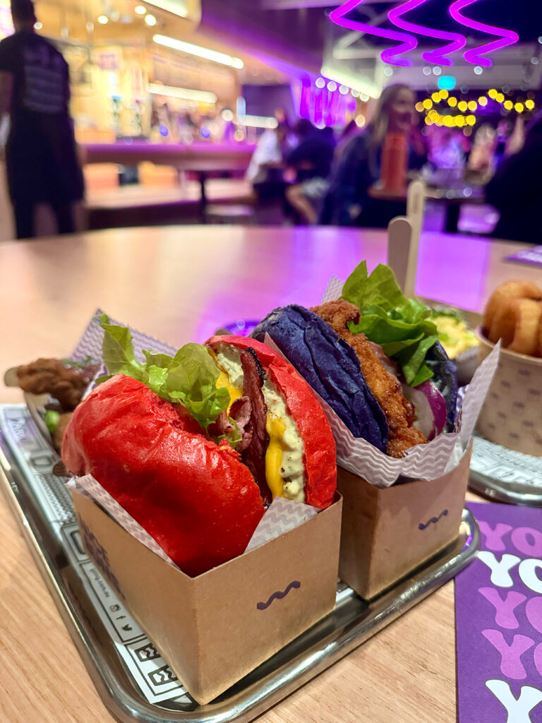 Two burgers sit on a wooden table, one with a pink bun one with a purple bun. There are purple neon lights above. 