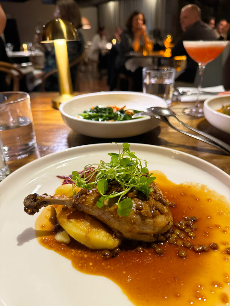 A white plate featuring a confit duck leg, mash potato and a lentil sauce with green garnish on top.