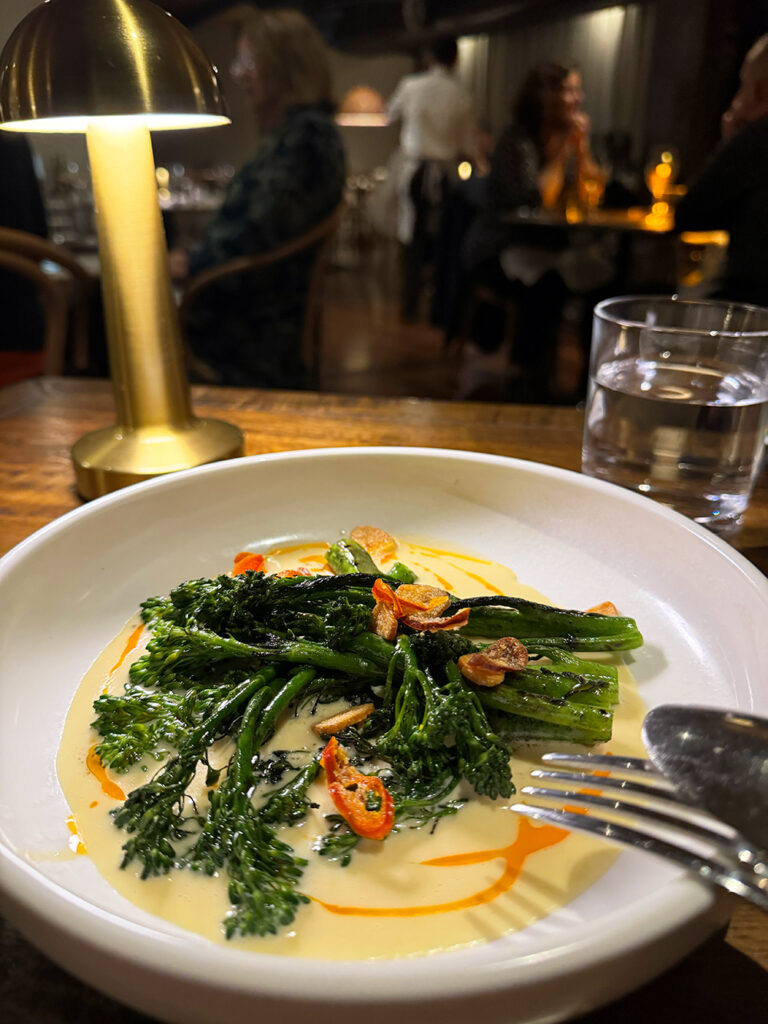 A white bowl contains chargrilled broccolini in a creamy gruyere cheese cause with chilli slices on top.