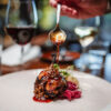A hand drizzles a sauce on top of a white plate featuring a confit duck dish.