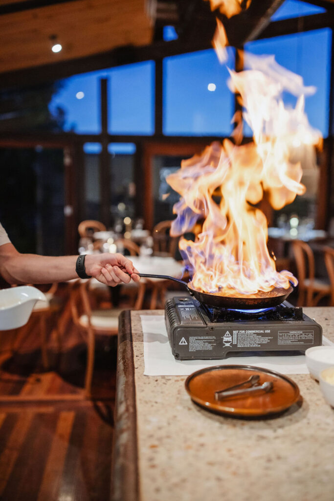 A hand holds a pan that is on fire with crepes suzette inside. 