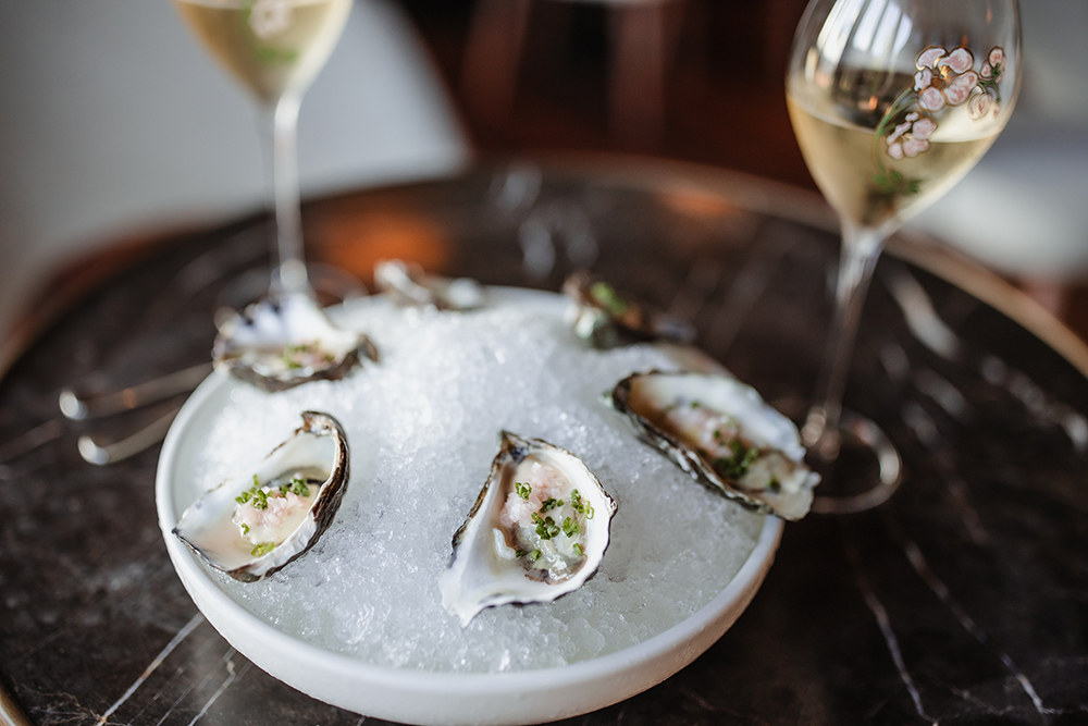 Two white wine glasses sit beside a platter with six rock oysters on a bed of salt.