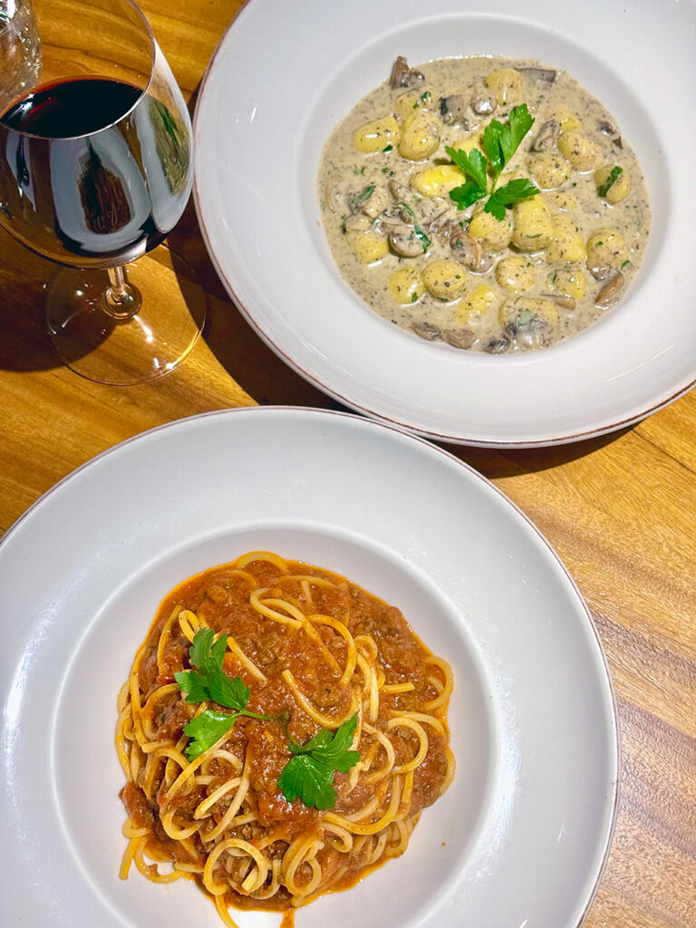 A bowl of spaghetti bolognaise and a creamy mushroom gnocchi sit on a wooden table beside a glass of red wine. 