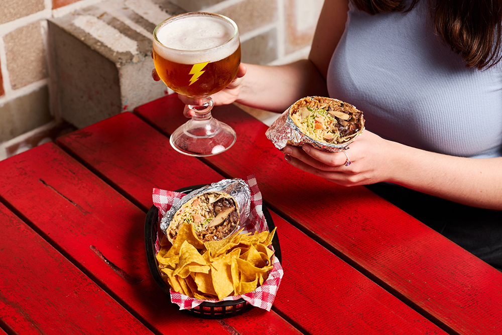 A woman's hands hold a burrito and beer, over a red table. The table also has a basket of corn chips and another burrito on it.