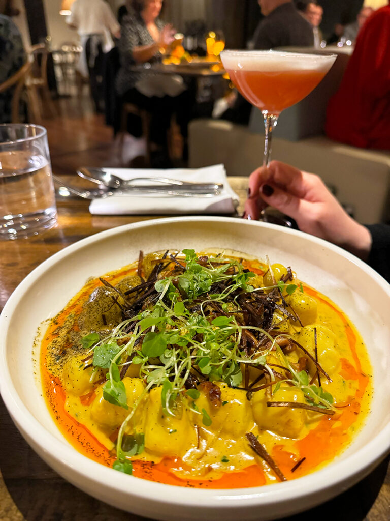 A while bowl featuring gnocchi with a yellow sauce and greens on top. A hand holds a martini glass in the background. 