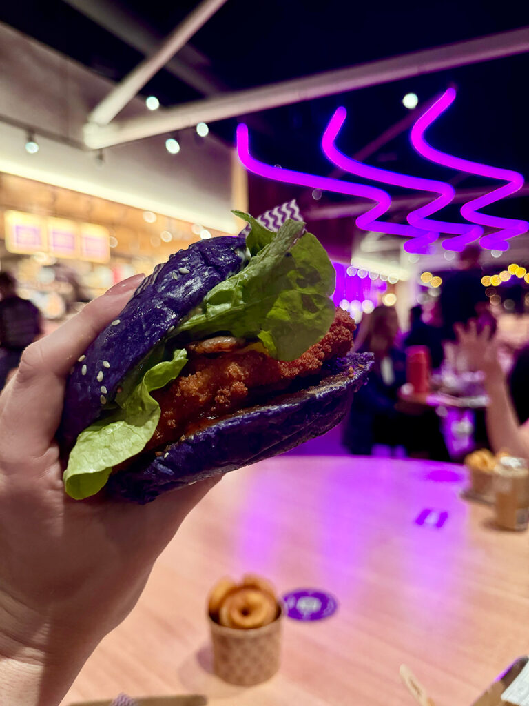 A woman's hand holds a chicken schnitzel burger with a purple bun up. There are purple neon lights in the background.