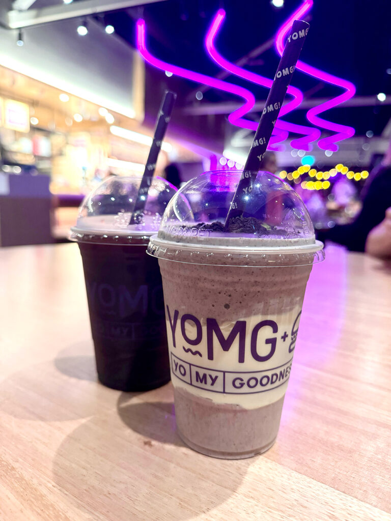 An OREO milkshake sits in front of a cola slushie on a wooden table. The brand YOMG is visible on the front of the cups.