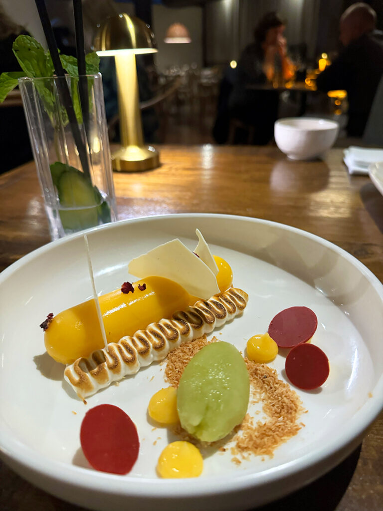 A white plate features a yellow tarte next to a squiggle of meringue, toasted coconut and sorbet. 