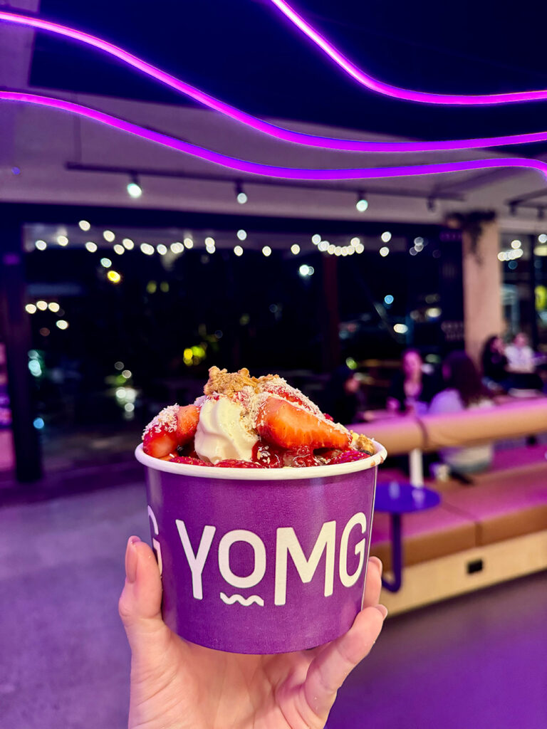 A woman holds up a purple cup with YOMG on the front, filled with chocolate and vanilla frozen yoghurt covered in strawberries and coconut.