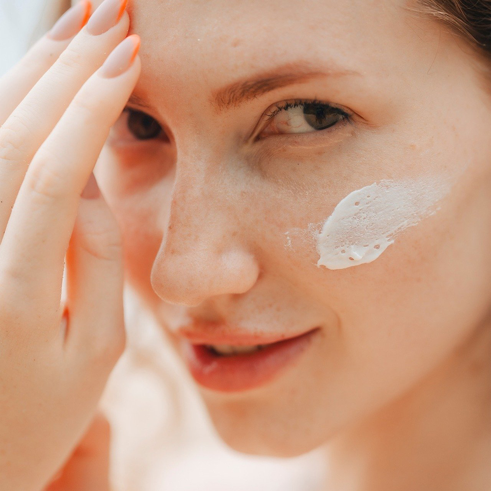 Close up on a woman's face. She has freckles, is putting on moisturiser and smiling at the camera.