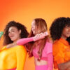 Three women in brightly coloured clothing pose with amika brand haircare products on an orange background