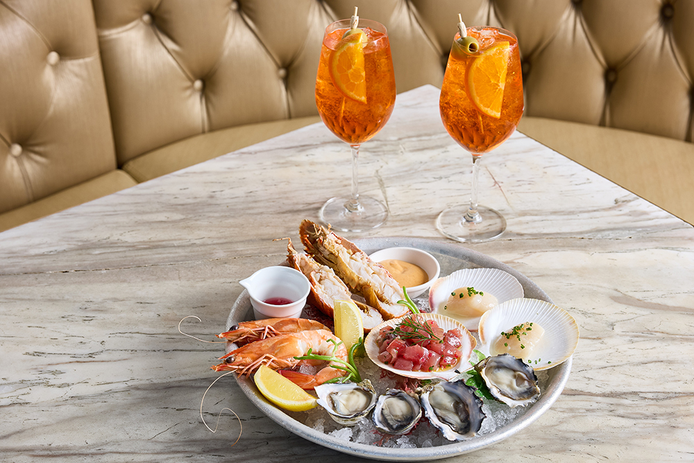 A seafood platter and two aperol spritzs sit on a marble table.