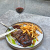A plate of steak frites and jus sits on a grey marble table, accompanied by a glass of wine.