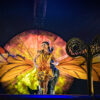 A woman with massive monarch butterfly wings stands behind a big key for Cirque du Soleil Luzia