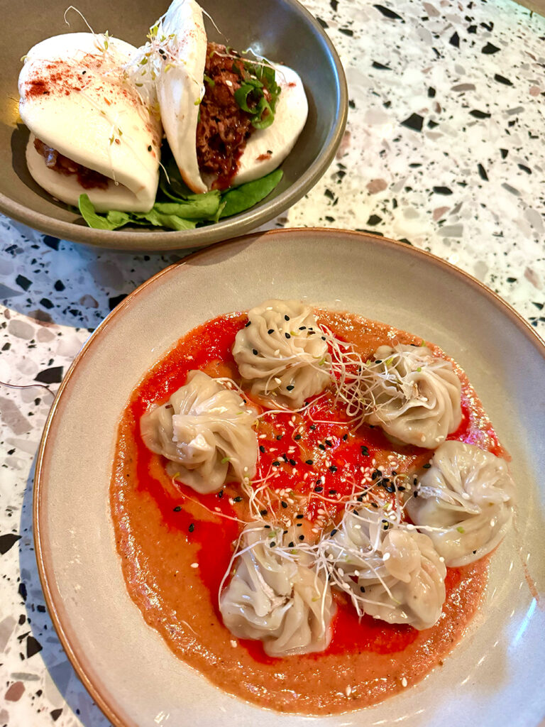 A plate of two duck bao buns sits alongside a plate of steamed momo dumplings in red sauce. Both sit on a terazzo table. 