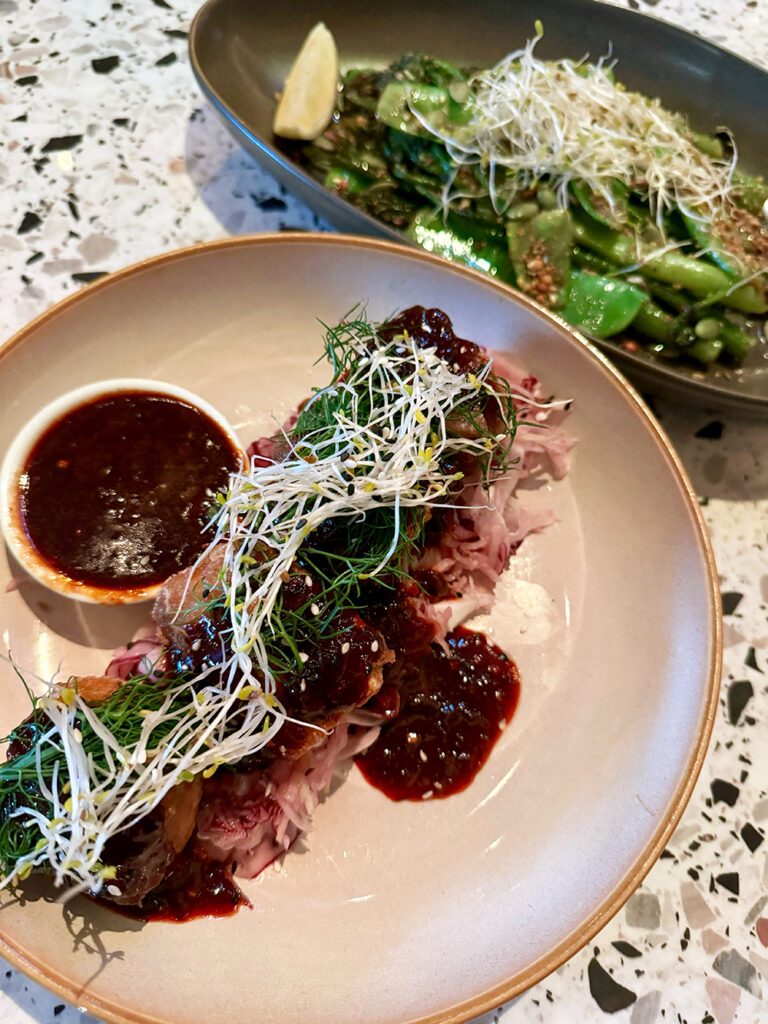 A plate of seasonal greens and a plate of pork belly sit on a terazzo table. 