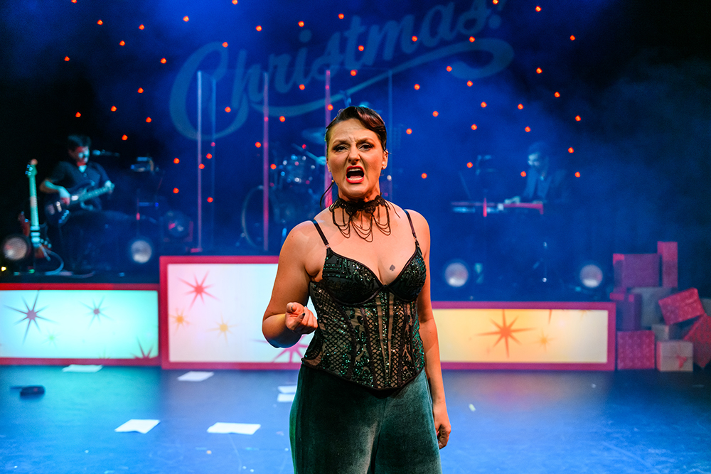 A woman in a green corset and green velvet pants points angrily at the camera while singing in front of the word Christmas. 