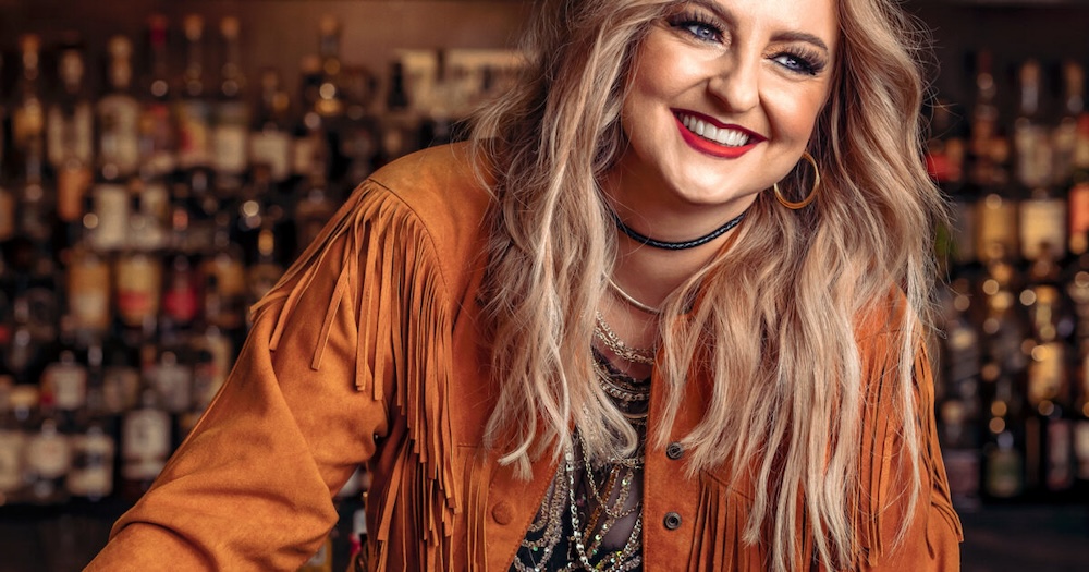 A blonde woman wearing a brown fringed jacket sits in front of a wall filled with bottles of spirits. 