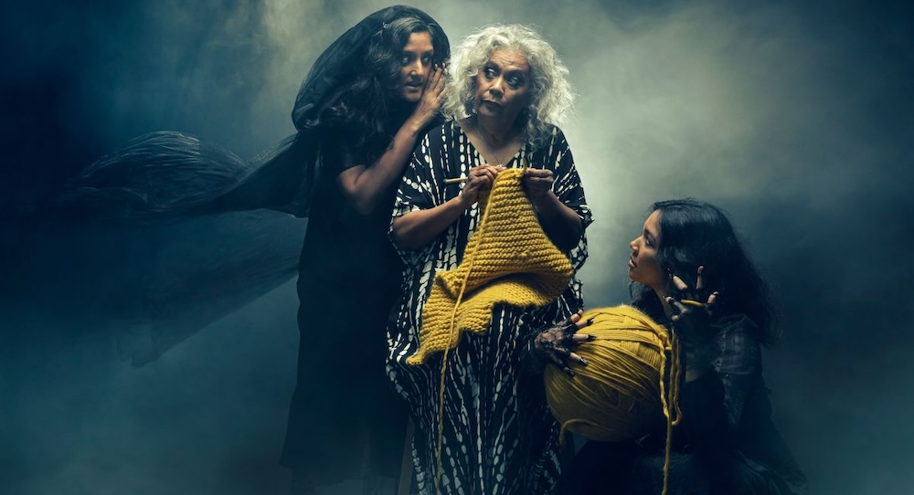 Three women dressed as witches whisper between each other while one in the centre knits with yellow yarn.