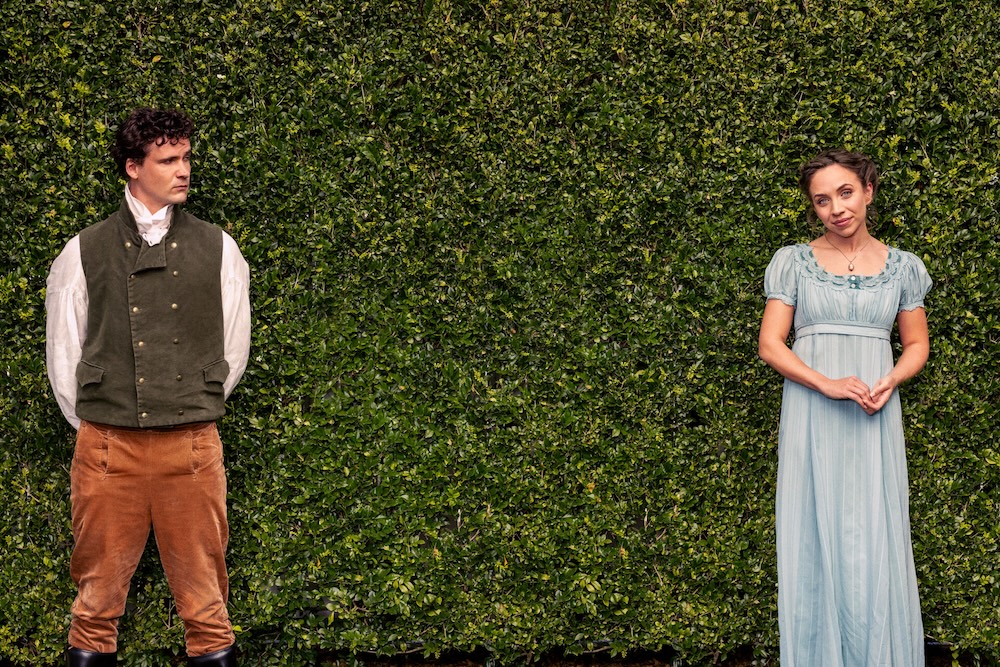 A man and a woman dressed in regency attire stand apart from each other in front of a hedge.