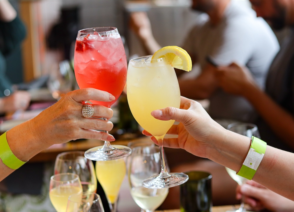 Two hands cheersing cocktails, one red and one yellow with a lemon wedge.