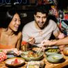 A man and woman with dark hair smile as they share japanese food on a wooden table