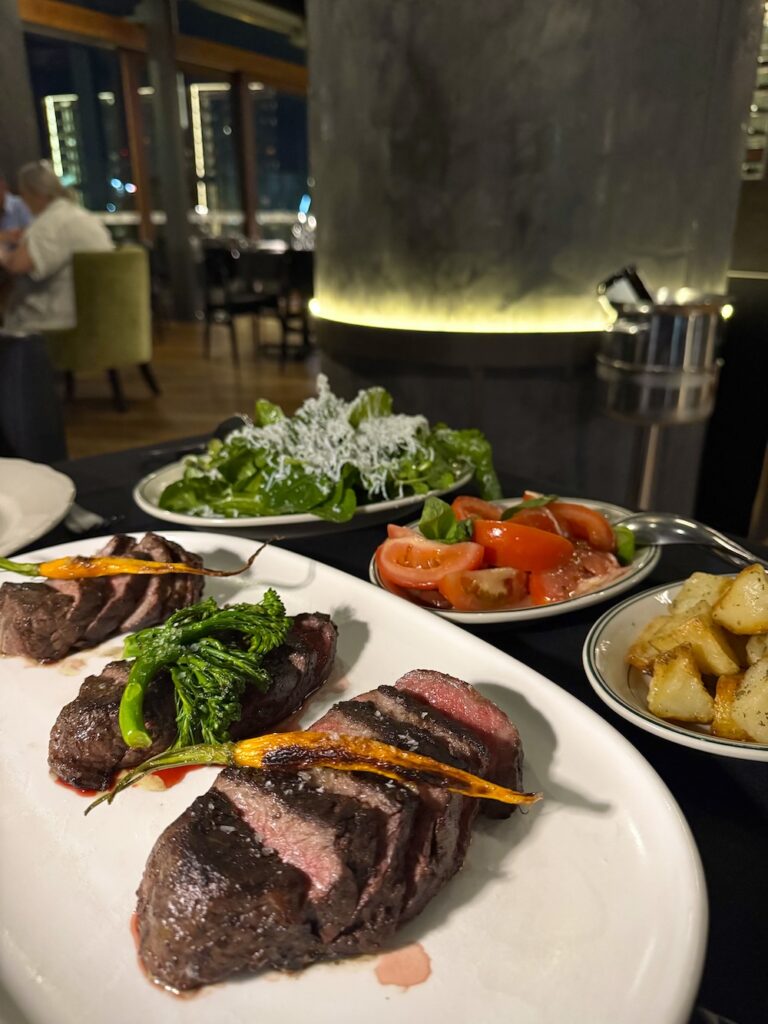 Three sliced steaks sit in the foreground, with salad, tomatoes and crispy potatoes in the background.