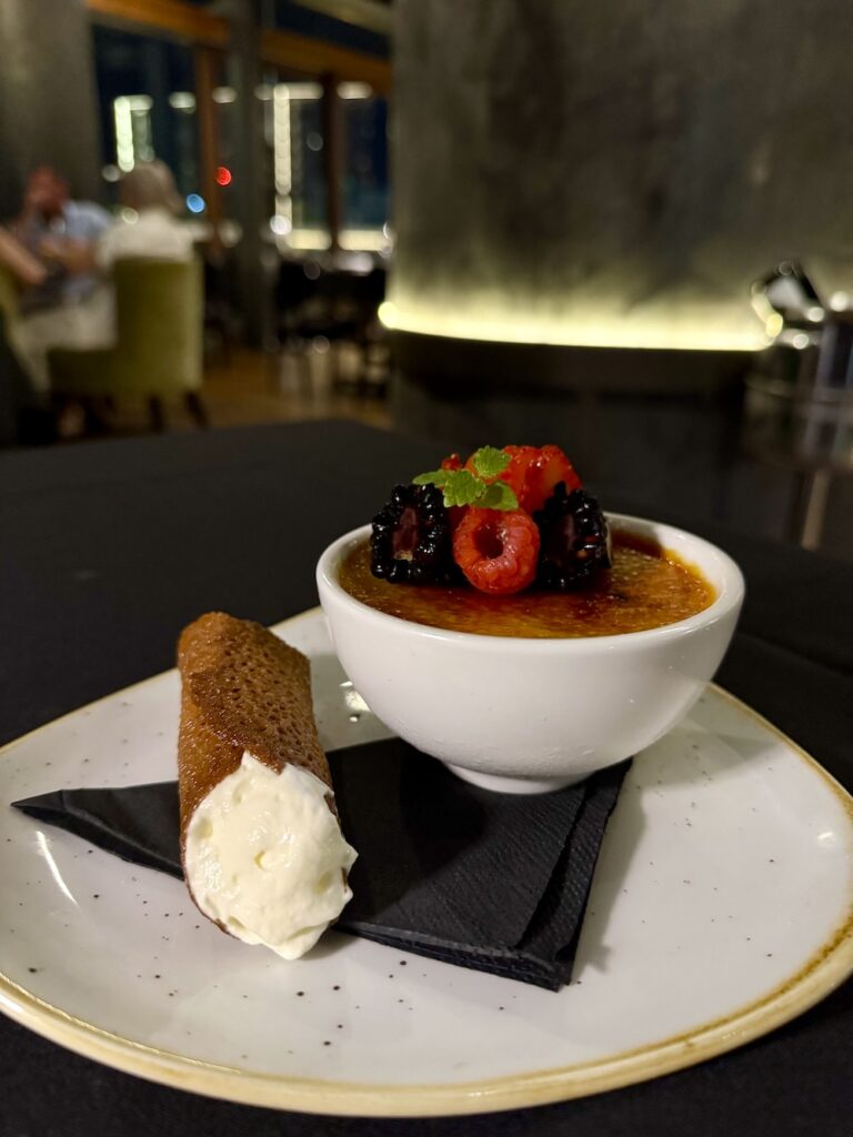 A creme brulee in a small bowl sits on a plate. Next to it is a brandy snap filled with chantilly cream.