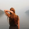 Woman with copper coloured hair with her back to the camera, staring out at a lake