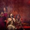 A woman sits on a red chaise with two see through women flanking her on the left and the right—ghosts