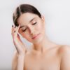 A brunette looks away from the camera as she touches her face. She appears with bare shoulders and clean skin