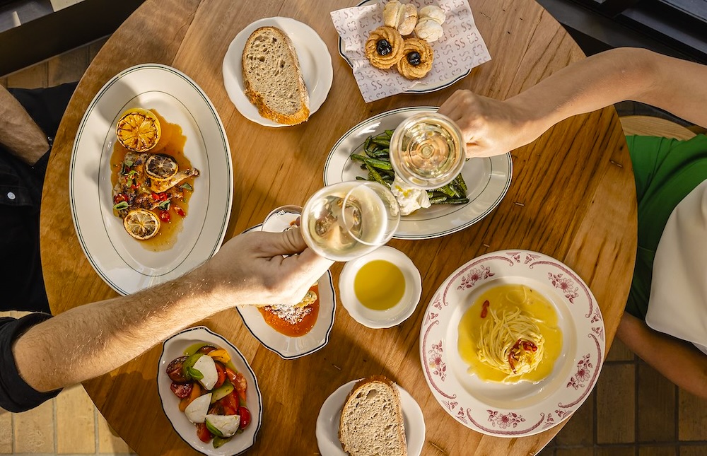 Experience an Italian feast at this hidden dining room at Settimo Two hands cheers over the top of a light wooden table filled with italian dishes at Settimo