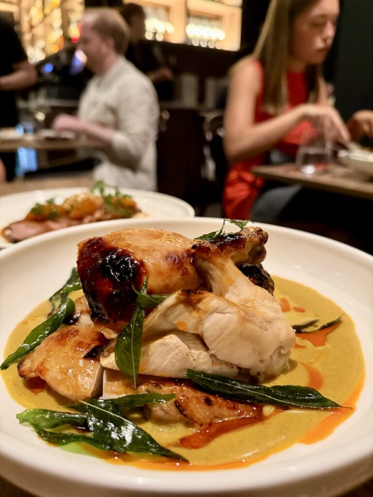 A white plate features several parts of chicken arranged on a yellow sauce bed with green leaves as a garnish at Monal dining