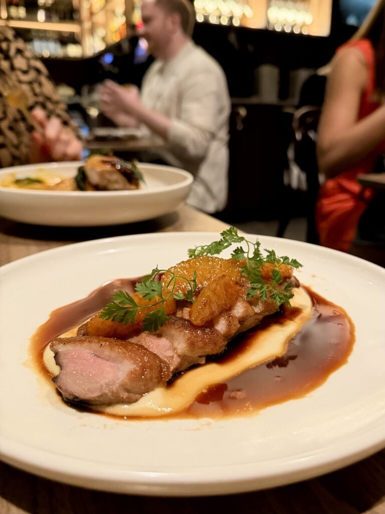A sliced duck breast with teriyaki orange slices sits on top of a silky brown sauxe on white plate. 