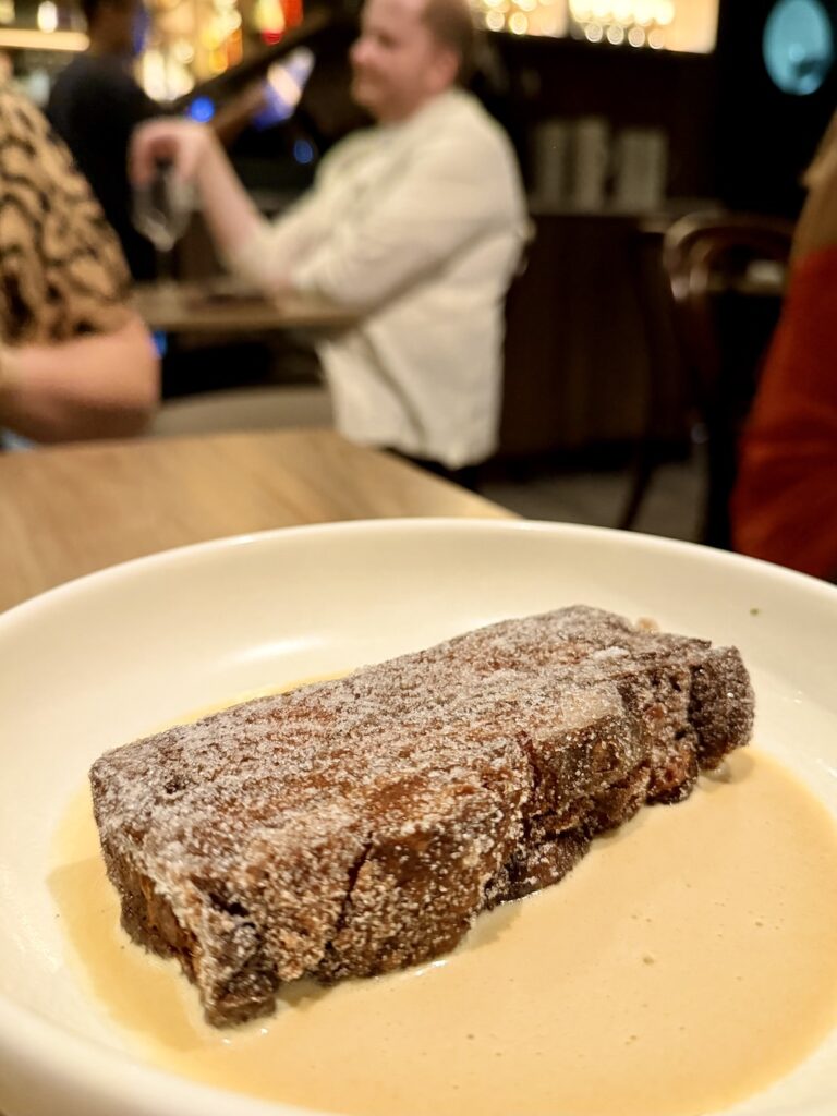 A crispy slice of fried bread and butter pudding coated with cinnamon sugar sits in a small pool of custard on a white place. 