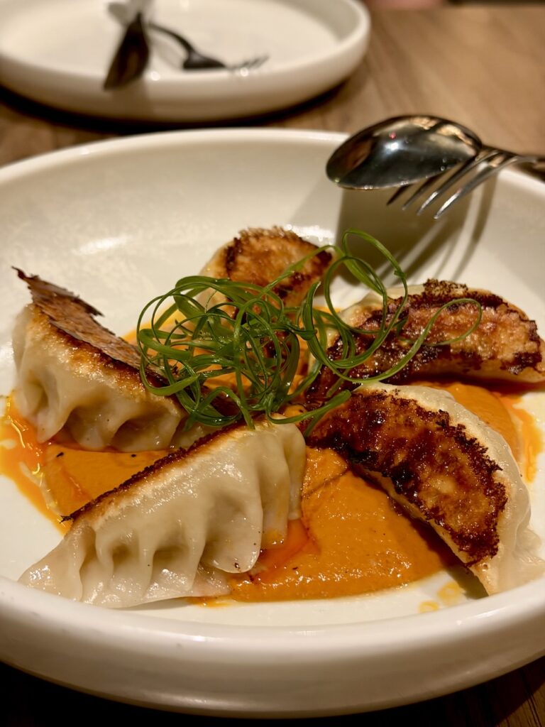 Five dumplings are arranged on a white plate in a star on a bed of orange sauce with green garnish at Monal Dining.