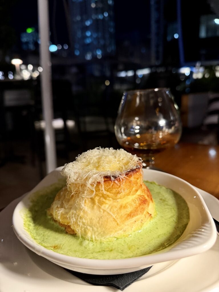 A gruyere souffle sits in a green sauce with a glass of cognac behind it at Pompette Brisbane 