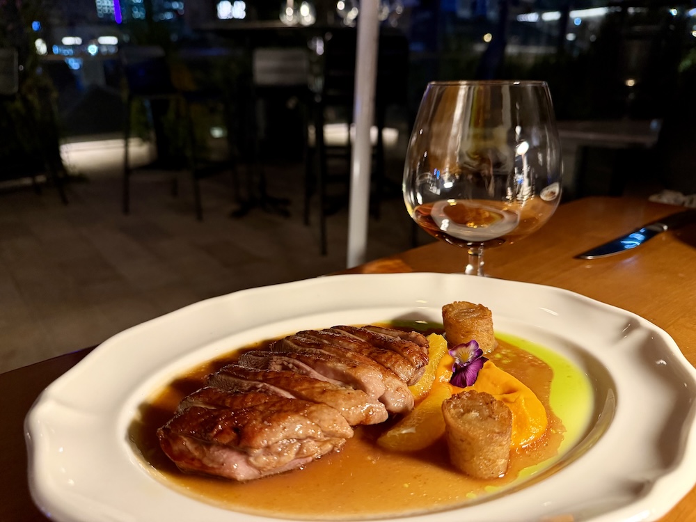 A sliced duck breast in a pool of a l'orange sauce sits on a white plate, with potato rilettes. A glass of cognac sits in the background at Pompette Brisbane