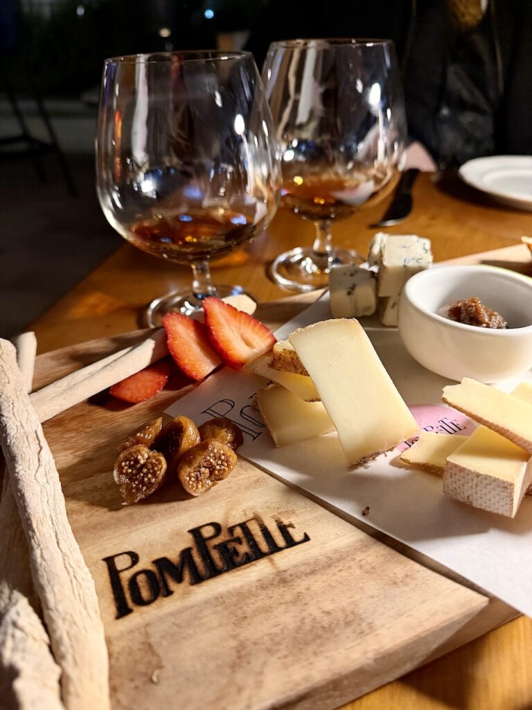 A selection of breads and cheeses sits on a wooden charcuterie board with Pompette burned into the board. 