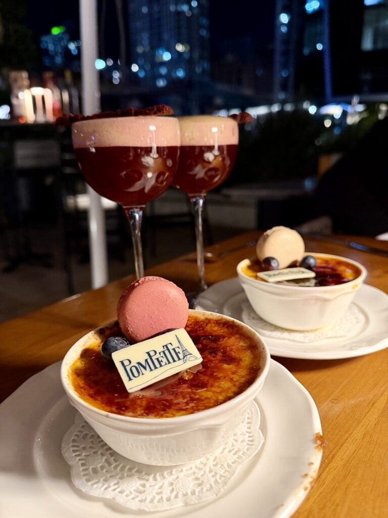 Two creme brulee's sit next to two raspberry cognac cocktails. Each creme brulee has a macaron and berries on the top as well as a white chocolate plaque with pompette printed on it
